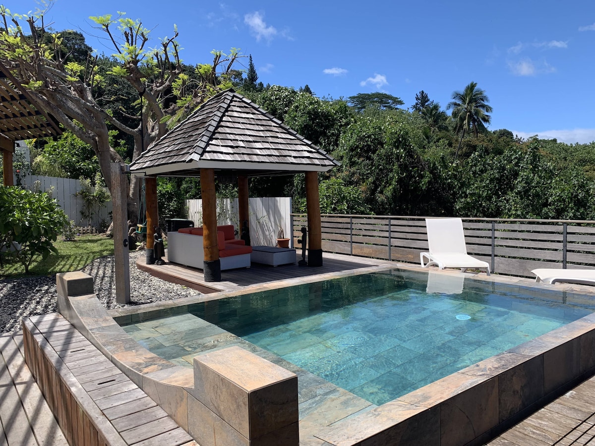 An outdoor living space is showcased, featuring a salt-water pool surrounded by wooden decking. A thatched-roof pavilion is visible, offering shaded seating with a red couch. Lush greenery and tropical trees provide a natural backdrop, enhancing the relaxed environment.