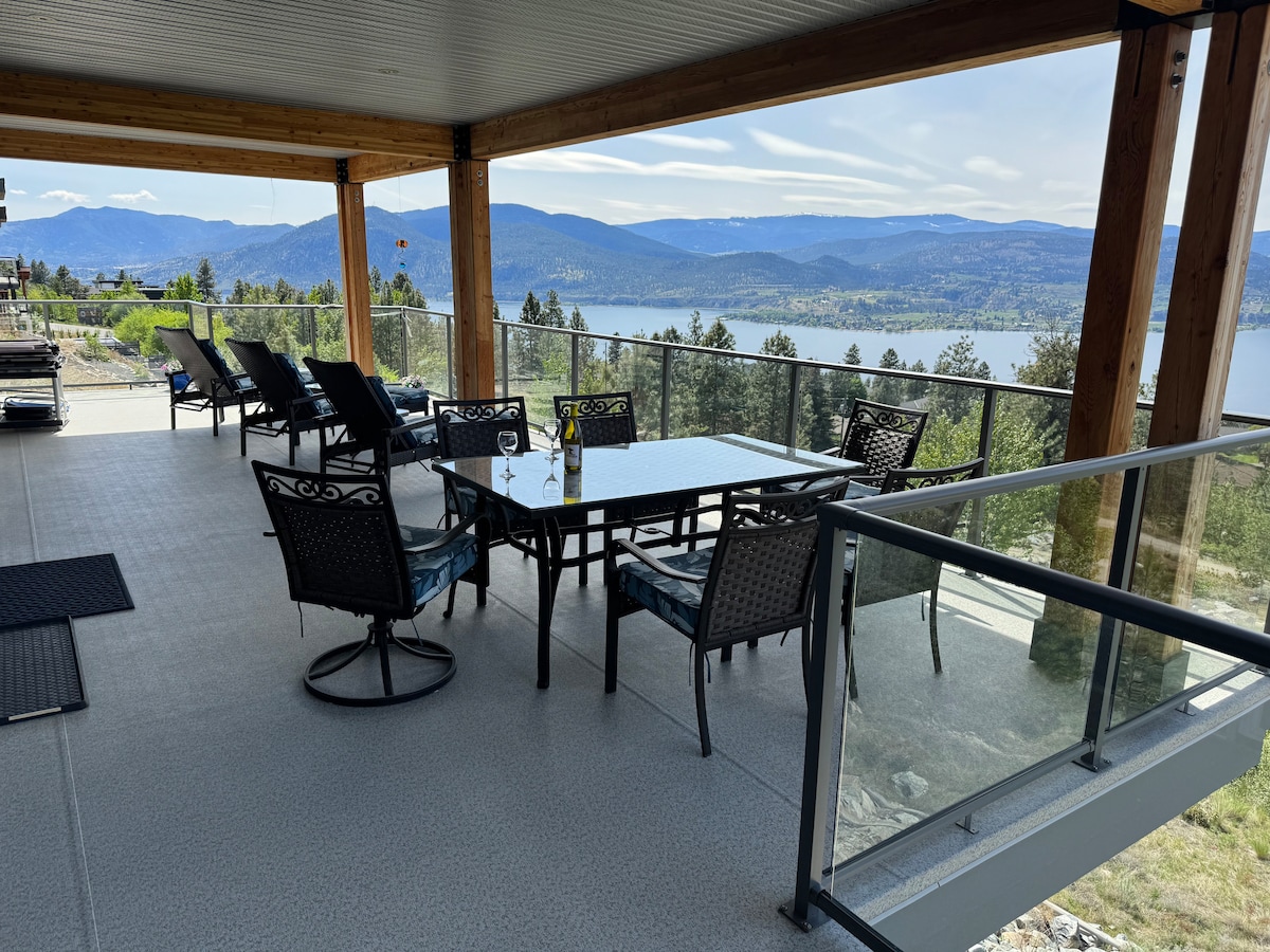 An expansive outdoor deck is furnished with a large dining table and several lounge chairs. A sweeping view of Lake Okanagan is visible through the railing, framed by distant mountains and lush greenery. The space is designed for relaxation and enjoying the scenery.