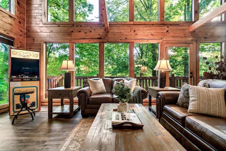 Gather in the cozy living room featuring a gas fireplace, PacMan multicade arcade game, Queen sleeper sofa, foosball table, Roku smart TV, and board games, all set against breathtaking outdoor views framed by expansive floor-to-ceiling windows