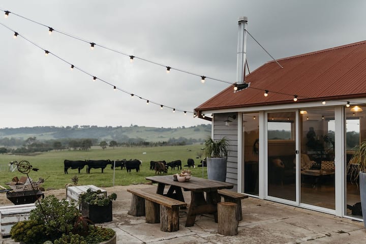  The image shows an idyllic countryside scene featuring an outdoor bathing area with a black freestanding tub and shower. The space is surrounded by lush greenery and grazing cows in a nearby field, creating a peaceful, rural atmosphere. String lights hang above the patio, adding charm to the setting. Rolling hills in the background enhance the scenic view, making this a perfect spot to relax and enjoy nature in a tranquil farm environment.