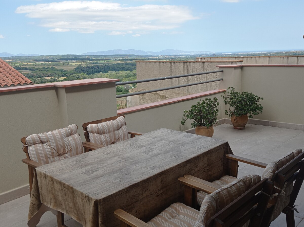 A private terrace is presented with a wooden dining table surrounded by four wooden chairs. Potted plants add a touch of greenery, while expansive views of the countryside and distant mountains are visible under a partly cloudy sky.