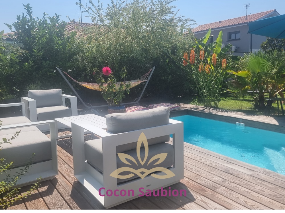 A serene outdoor space features a wooden deck with two modern armchairs positioned around a low table. A pool reflects the sunlight, surrounded by lush greenery and vibrant flowers. A hammock is visible in the background, enhancing the peaceful ambiance.