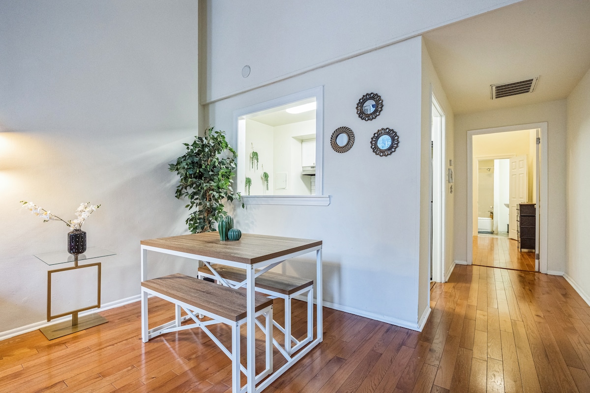 A dining area features a wooden table with bench seating and a decorative plant. A doorway leads to a bright kitchen area, while mirrors are displayed on the wall, reflecting natural light. A hallway is visible, providing access to additional rooms.