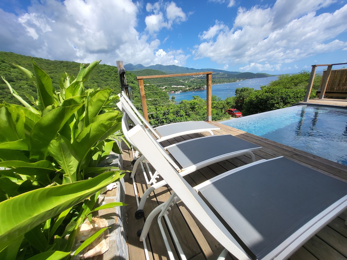 Lounge chairs are positioned by the infinity pool, with a view of the lush green hills and the shimmering blue waters of the bay. The scene captures clear skies scattered with clouds, enhancing the tranquil outdoor atmosphere.