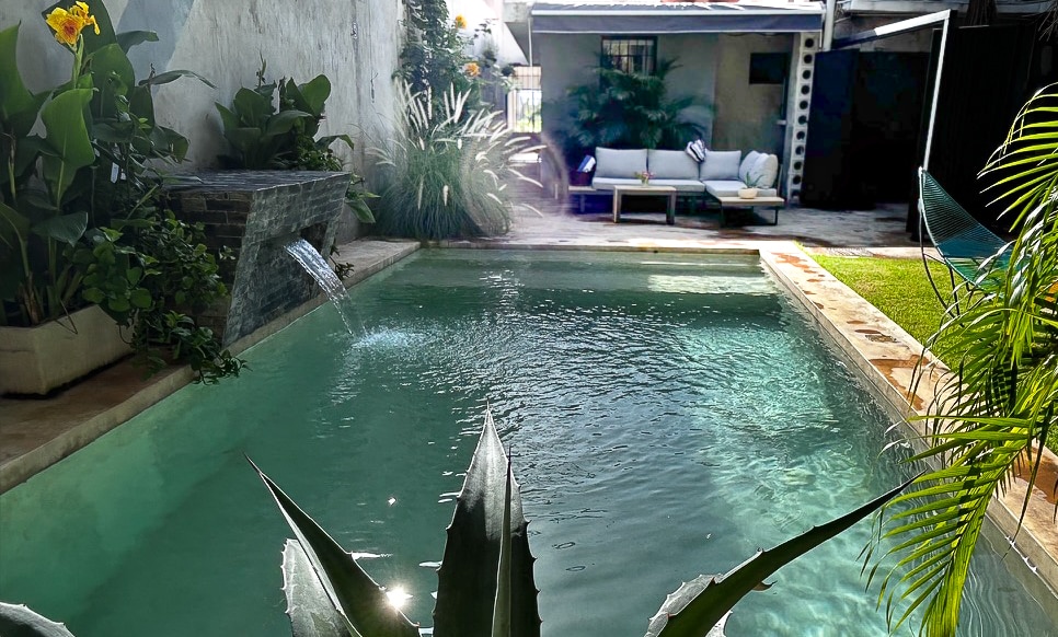 A tranquil pool is surrounded by lush greenery, with a gentle fountain feature adding to the serene environment. A comfortable outdoor seating area is visible in the background, providing a space for relaxation. Sunlight reflects off the water's surface, enhancing the peaceful setting.