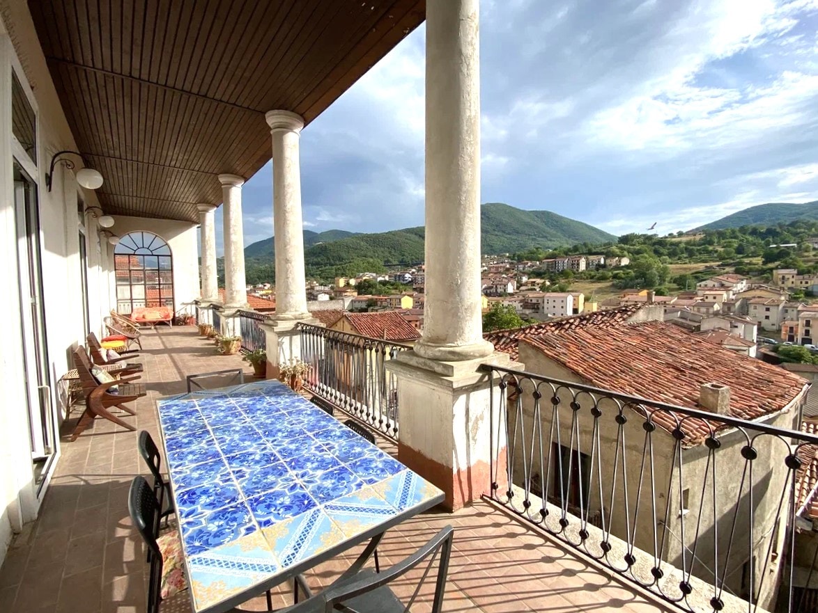 A spacious terrace is presented, featuring tall columns and a decorative tiled table. Seating options include chairs arranged for outdoor enjoyment, with panoramic views of the surrounding mountains and the historic town below. Natural light enhances the inviting ambiance.