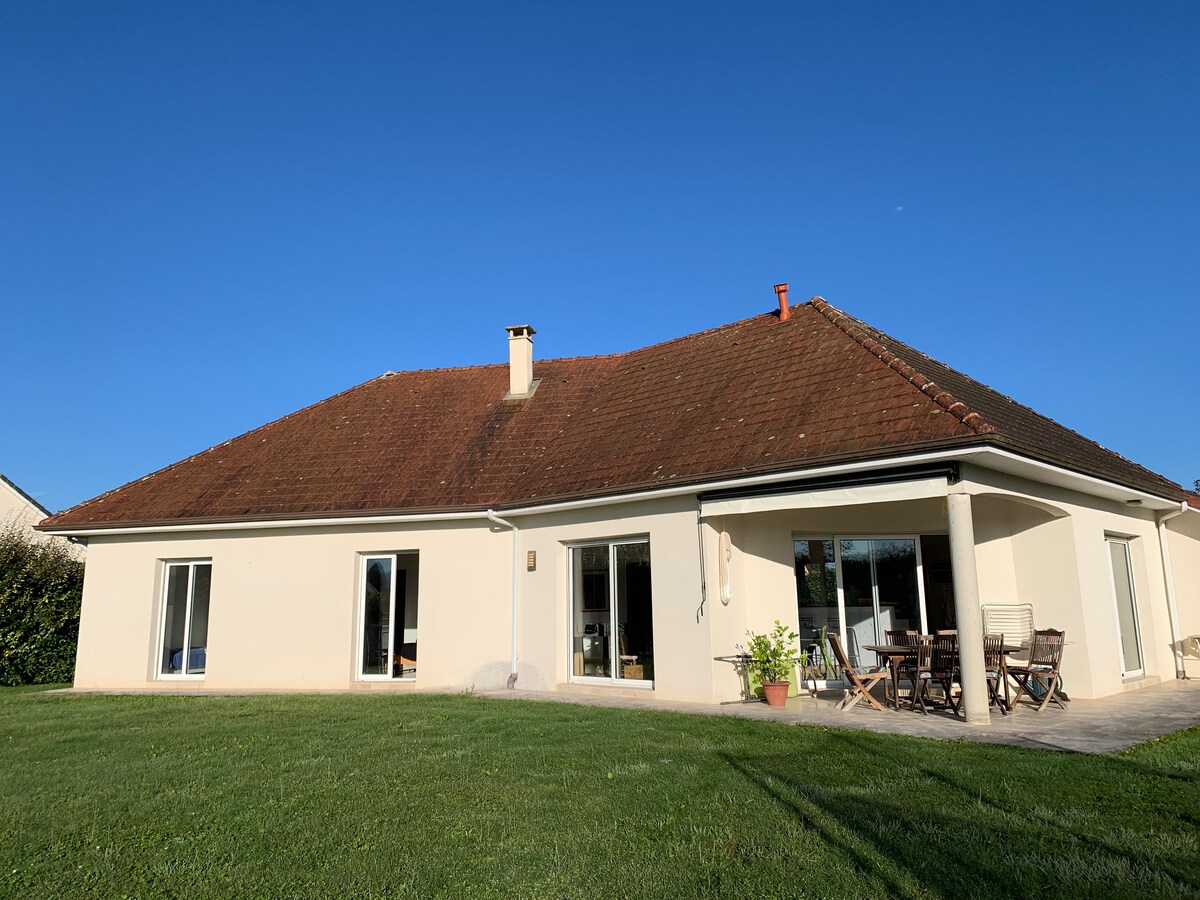 A single-story house is set against a clear blue sky, featuring large glass doors that provide easy access to the outdoor space. A well-maintained lawn surrounds the property, with a patio area furnished with a wooden dining table and chairs for outdoor gatherings.