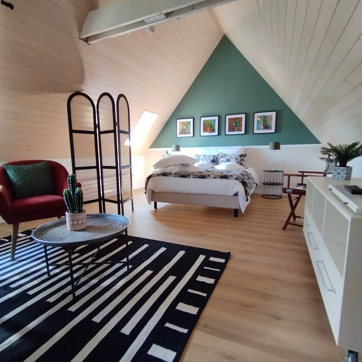 The spacious room features a sloped ceiling with wooden paneling. A large bed is situated near a window, complemented by framed artwork on the green wall. A cozy armchair and a room divider add comfort, while a patterned rug enhances the warm atmosphere of the space.