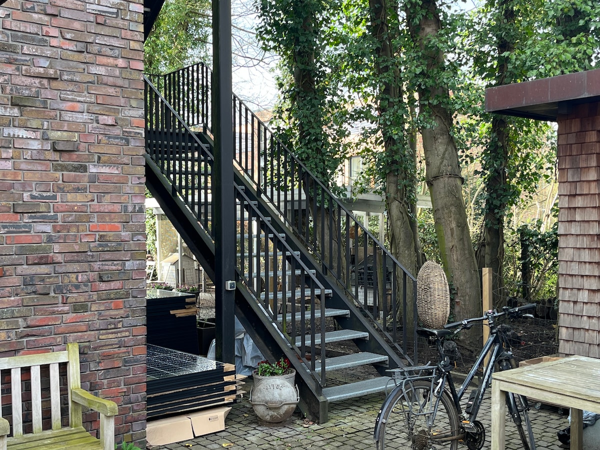 An exterior view of a black, metal staircase leading to an upper level, surrounded by greenery. A bicycle is parked nearby, and several steps are visible, along with a small seating area to one side.