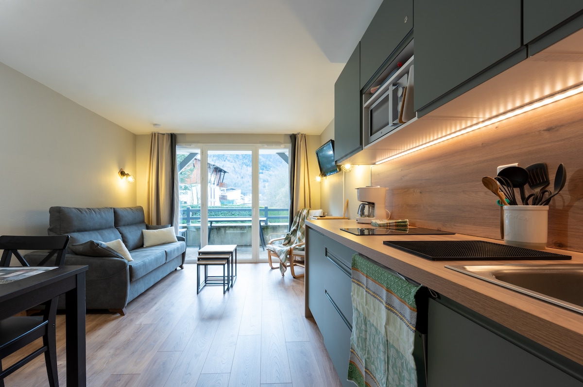 A bright studio is depicted, featuring a cozy seating area with a gray sofa and a small wooden table. A kitchenette with modern appliances and a sink is visible, complemented by warm lighting underneath. Large glass doors open to a balcony with mountain views.