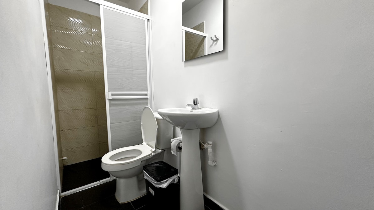 A clean and functional bathroom features a white toilet and a pedestal sink with a mirror above. The space is illuminated by soft lighting, and a wastebasket is positioned beside the sink. The walls are painted in a neutral tone, enhancing the room's brightness.
