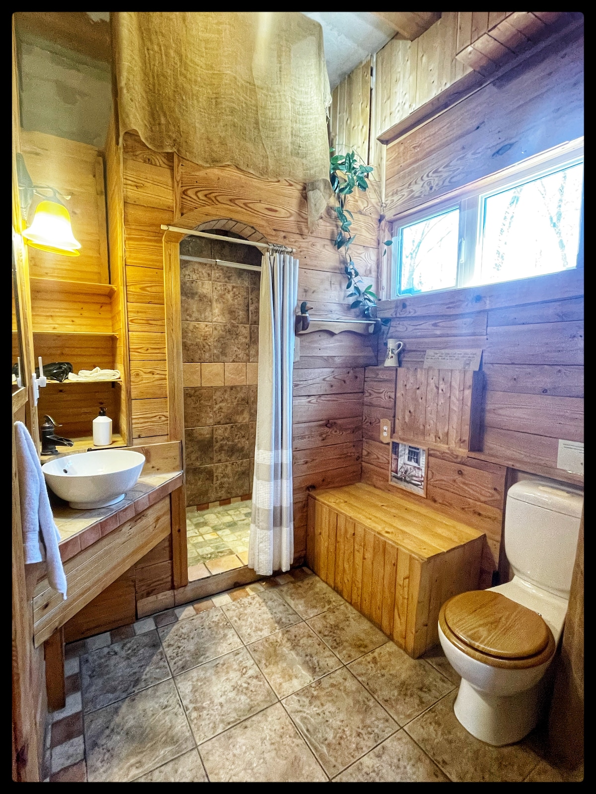 A wooden-themed bathroom features a spacious shower with textured tiles and a glass door. Natural light filters through a window, illuminating a vessel sink on a wooden vanity. An organized shelf holds toiletries, while a wood storage bench provides additional space.