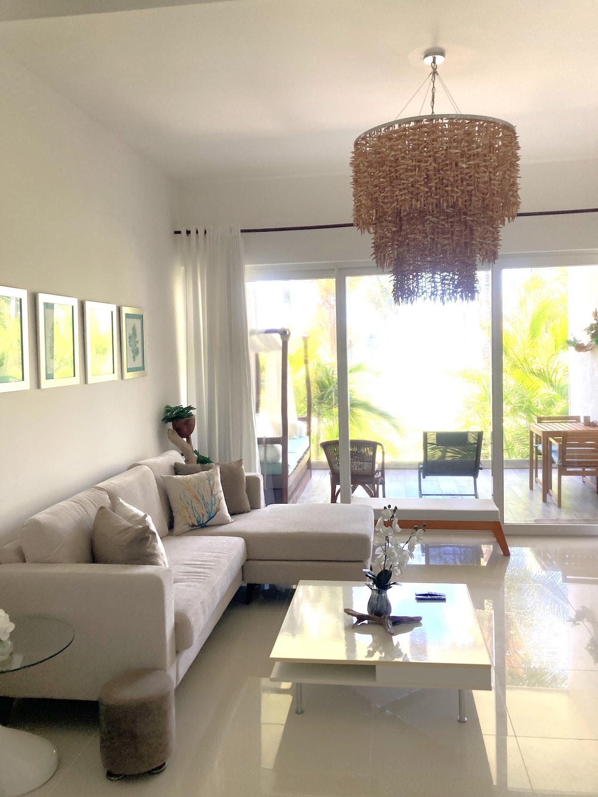 A well-appointed living area features a light-colored sectional sofa complemented by decorative cushions. A coffee table sits in front, with a small decorative plant. Large glass doors provide a view of the patio and palm trees, enhancing the bright and airy ambiance.