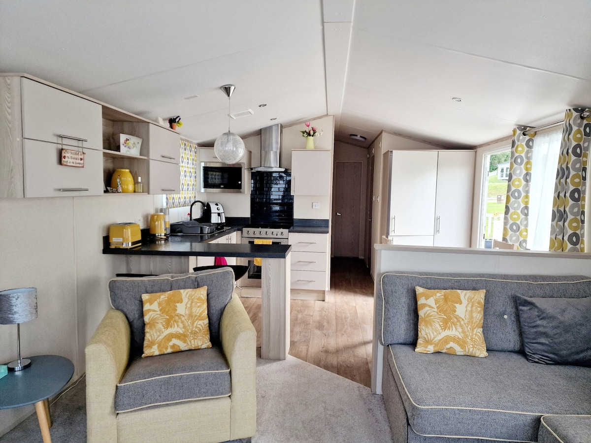 An open-plan living area showcases a modern kitchen and a cozy seating arrangement. Bright yellow accents are found in the kitchenware and throw pillows, complementing the soft grey upholstery of the sofas. The space is illuminated by natural light filtering through patterned curtains.