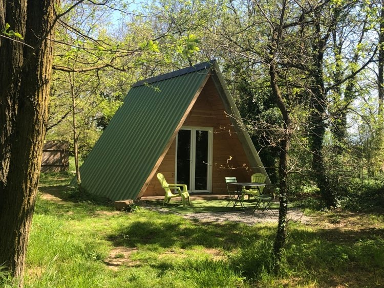 A charming A-frame cabin is surrounded by lush greenery, with large glass doors providing access to the outdoor area. Two green chairs and a small table sit on a grassy patch in front, inviting relaxation amid the natural setting.