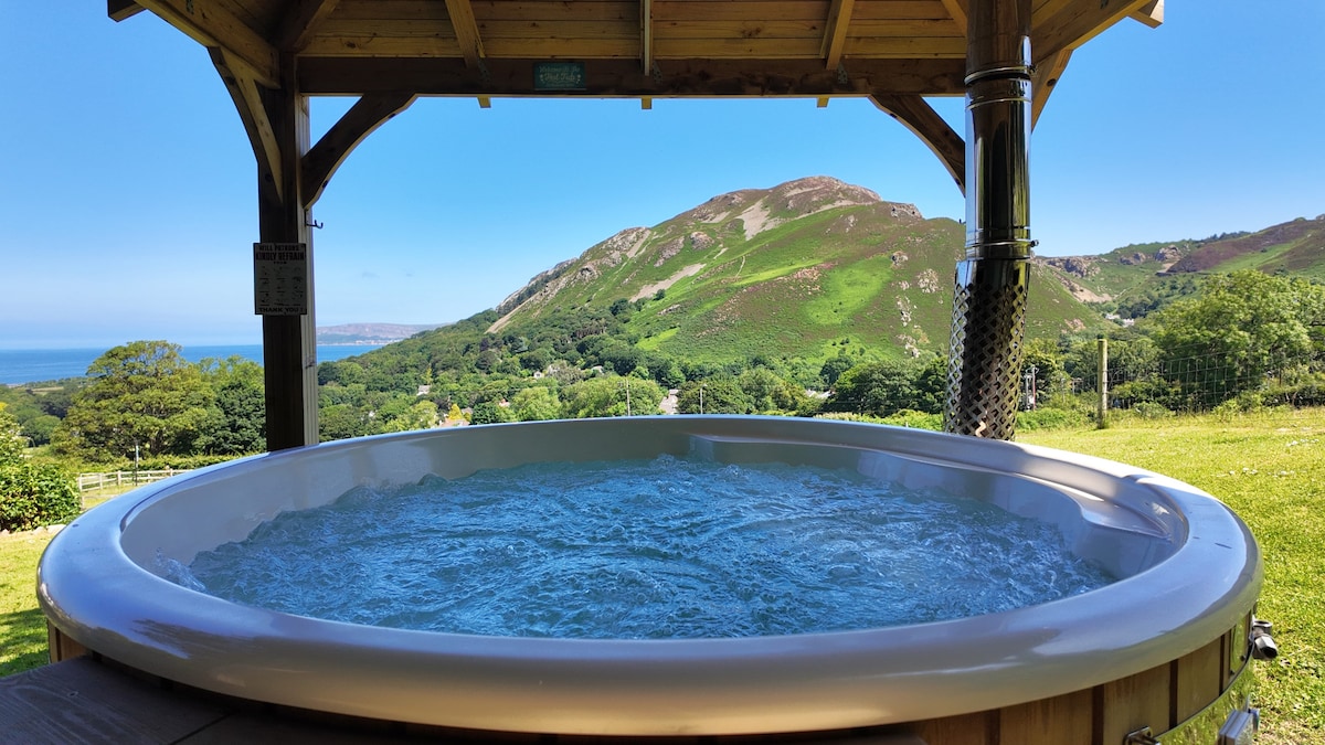 A log burning hot tub is showcased within a wooden gazebo, surrounded by a lush green landscape and hills in the background. The clear blue sky and sea are visible, emphasizing the tranquil setting for relaxation.