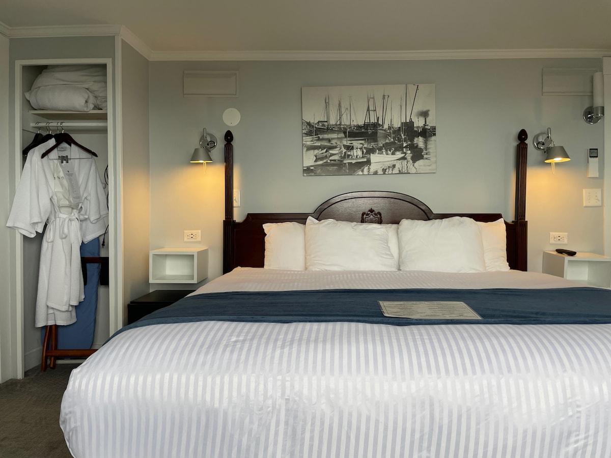 A spacious bedroom features a king bed with white linens and a blue runner. Flanking the bed are two nightstands with lamps. A closet with hanging robes is visible on the left, and a maritime-themed artwork adorns the wall above the bed.