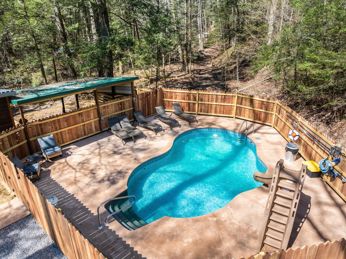The outdoor pool area is surrounded by a wooden fence, featuring a spacious, uniquely-shaped pool. Lounge chairs are positioned along the poolside, and a shaded structure offers a seating area nearby. The natural forest backdrop enhances the serenity of the location.