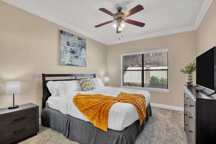 Dormitorio con una cama tamaño king con detalles en amarillo, muebles de madera oscura, un televisor sobre un tocador y una gran ventana que proporciona luz natural.