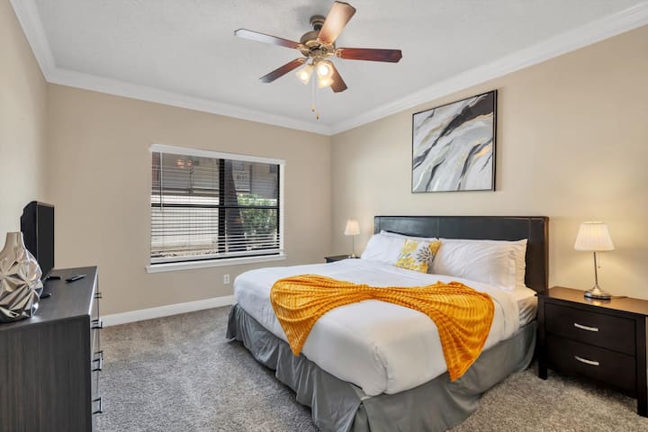 Dormitorio con cama tamaño king con detalles amarillos, muebles de madera oscura, paredes neutras, ventilador de techo y ventana con persianas.