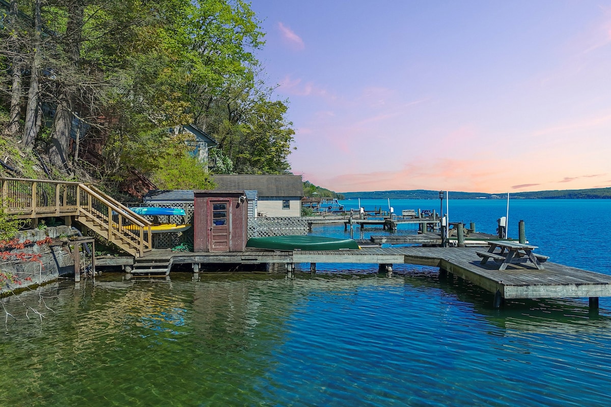 A waterfront area is captured with a private dock extending into the clear waters of Cayuga Lake. A picnic table is visible on the dock, alongside a storage shed. The surrounding greenery complements the tranquil setting as the vibrant sky reflects over the lake.