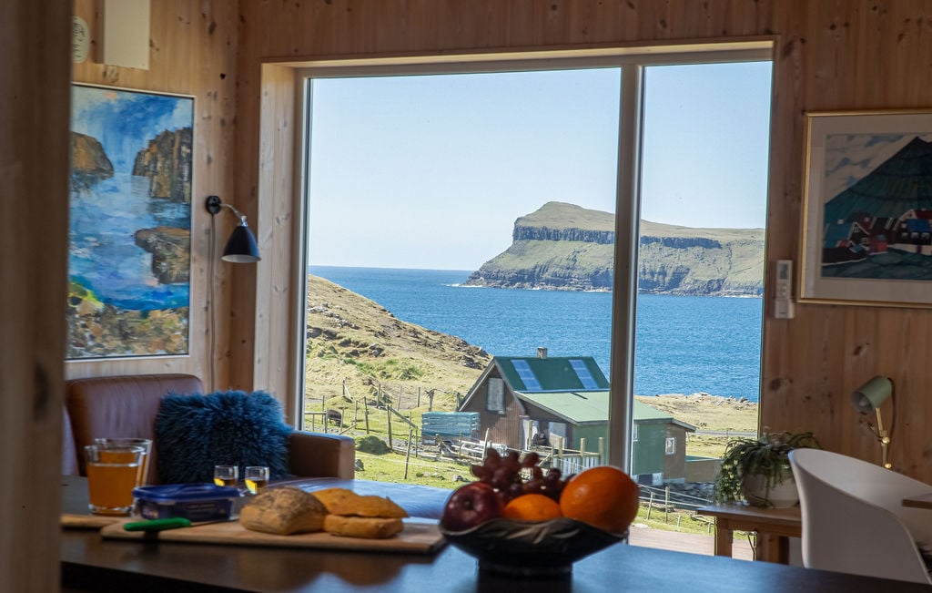 A large window frames a coastal view, highlighting expansive blue waters and nearby cliffs. Inside, a dining area is visible with a table set for breakfast, featuring fruits, pastries, and beverages. Nearby, colorful artwork adds a touch of character to the wood-paneled walls.