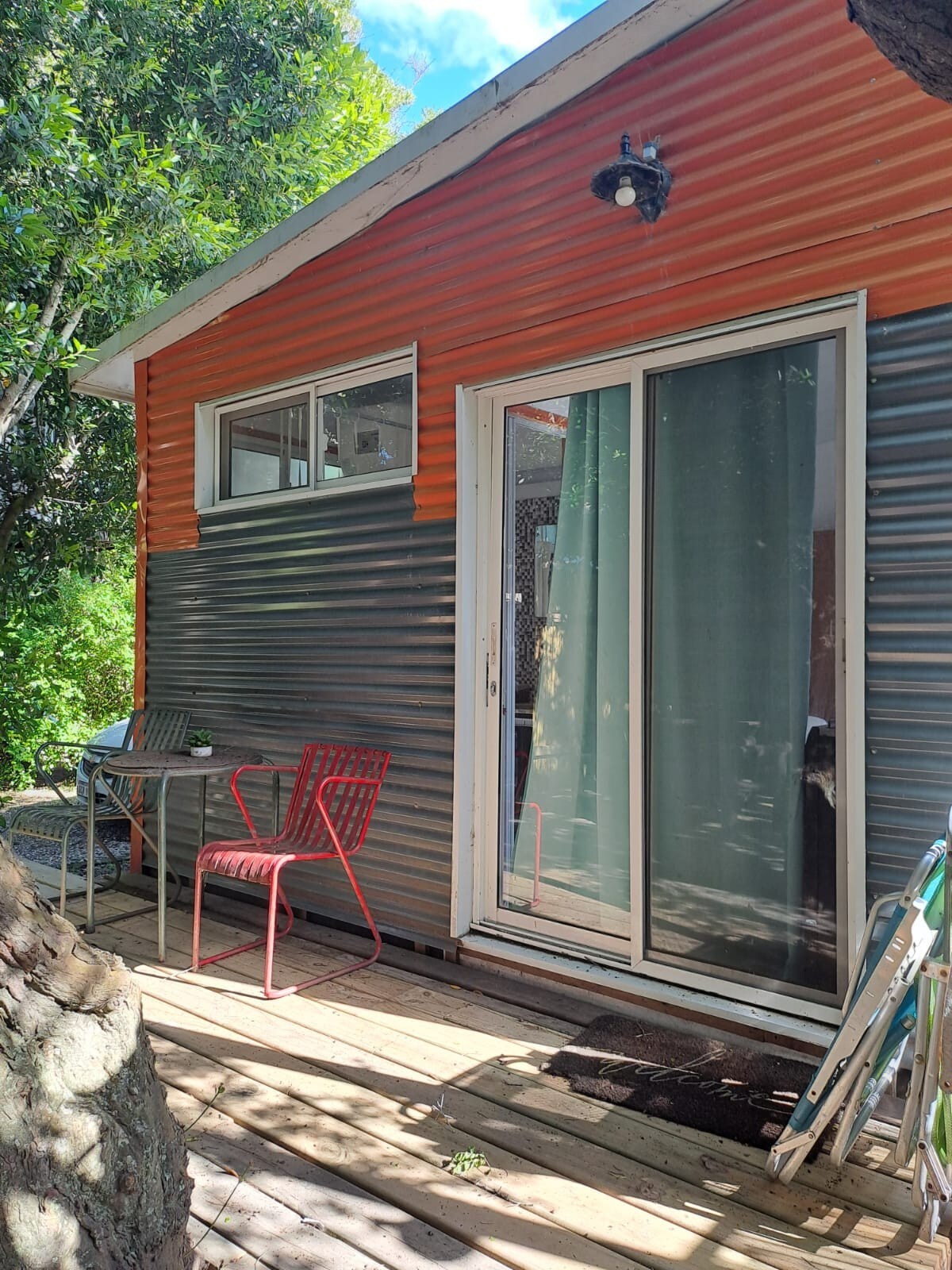 The exterior of a cabin is shown, featuring a combination of orange and gray corrugated metal walls. A wooden deck is visible with two red chairs and a small table. Glass sliding doors provide access to the interior, surrounded by lush greenery.
