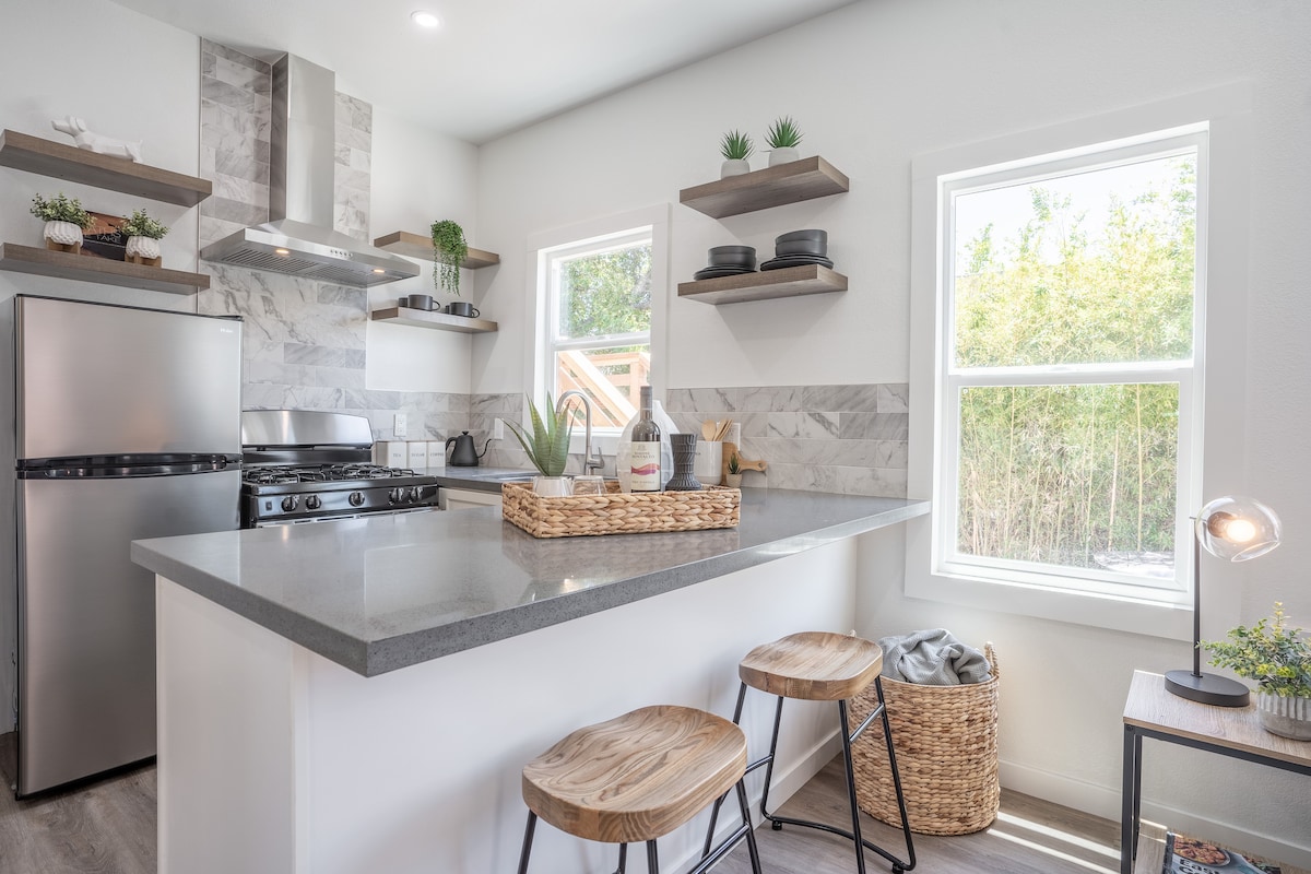 An inviting kitchen space features modern stainless steel appliances, including a refrigerator and a gas stove. A gray countertop supports a decorative tray and kitchen essentials. The room is brightened by natural light coming through two windows, with open shelves showcasing plants and dishware.