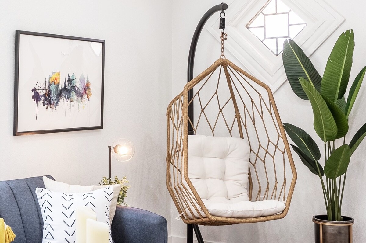 A cozy corner is highlighted by a unique hanging chair with a cushioned seat, resting within a modern frame. A vibrant piece of art adorns the wall while a potted plant adds a touch of greenery, complementing the light-filled space.