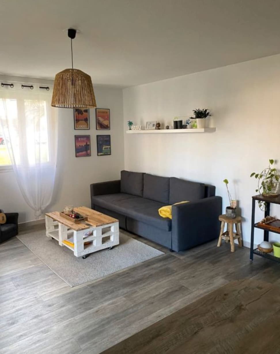 A cozy living area features a gray sofa complemented by a wooden coffee table, crafted from pallets. A light-colored area rug lies beneath, while framed artworks adorn the white walls. Natural light filters through sheer curtains, and decorative plants are positioned on a shelf and side table.