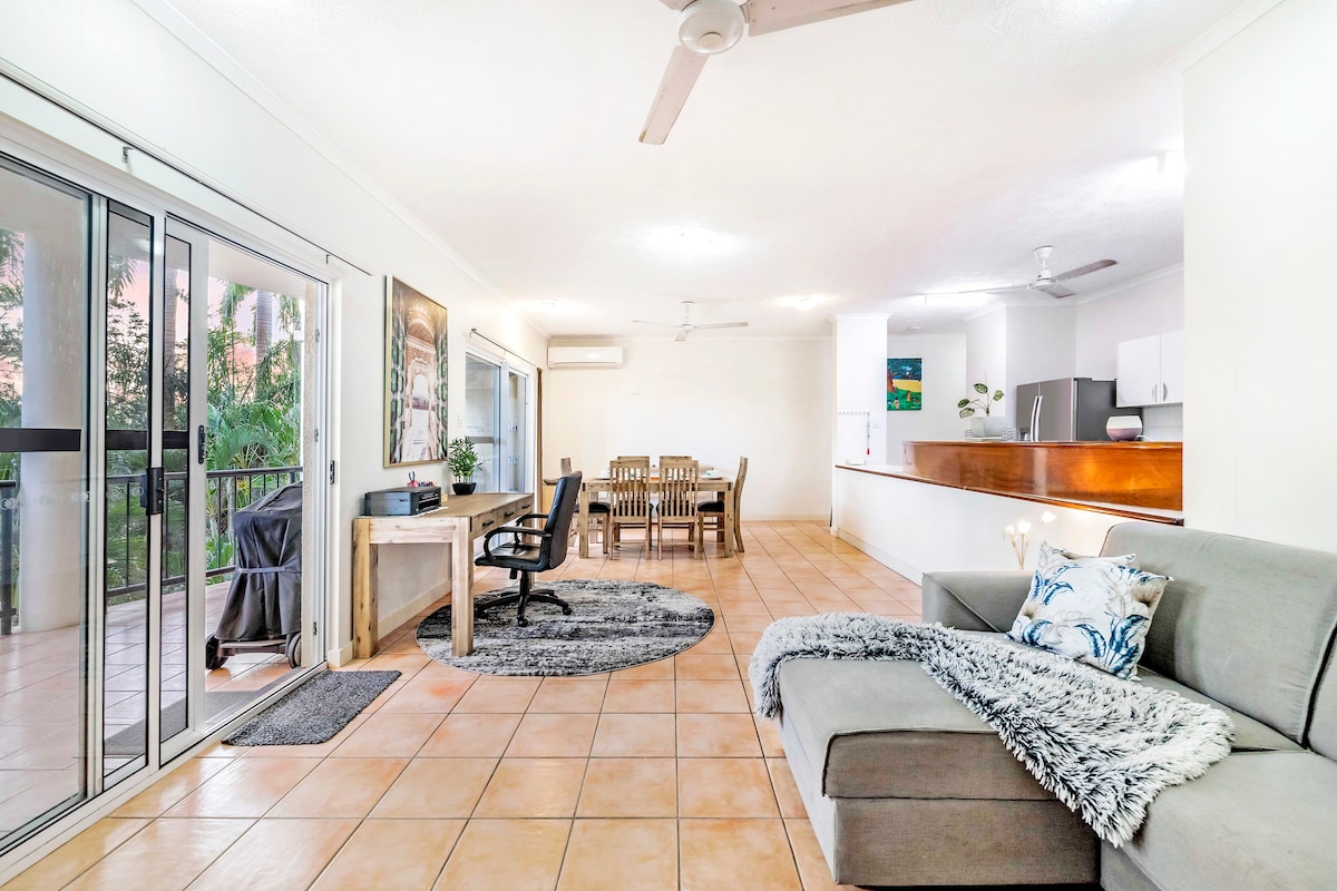 A spacious living area features a comfortable sofa and a large area rug. A wooden dining table sits to the right, accommodating multiple guests. Sliding glass doors open to a balcony, allowing natural light and fresh air to flood the space.