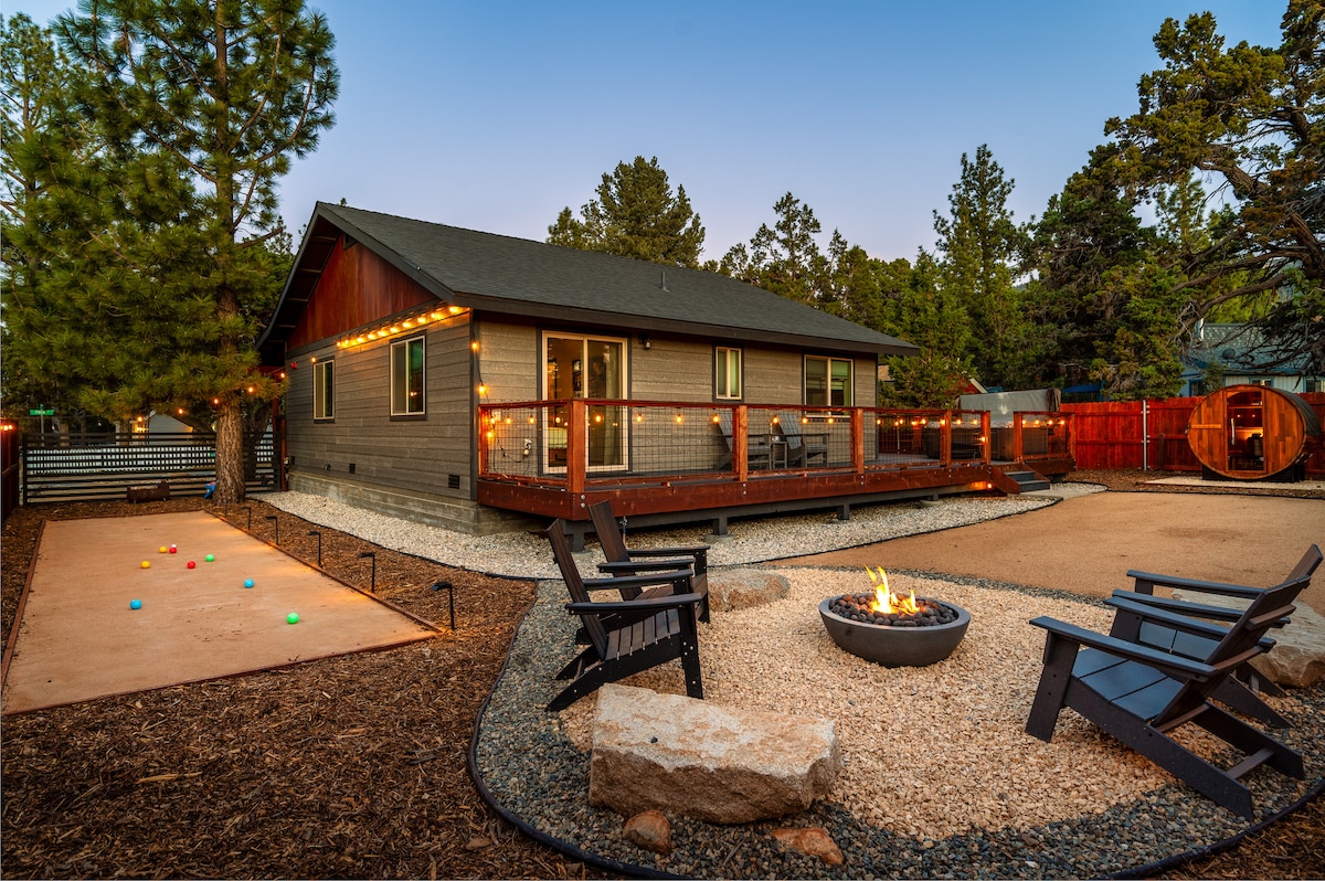 The outdoor area features a spacious deck with string lights illuminating the surroundings. A fire pit is centered between four black wooden chairs, providing a cozy gathering spot. A gravel pathway leads to a bocce ball court, enhancing the recreational space in the scenic backyard.