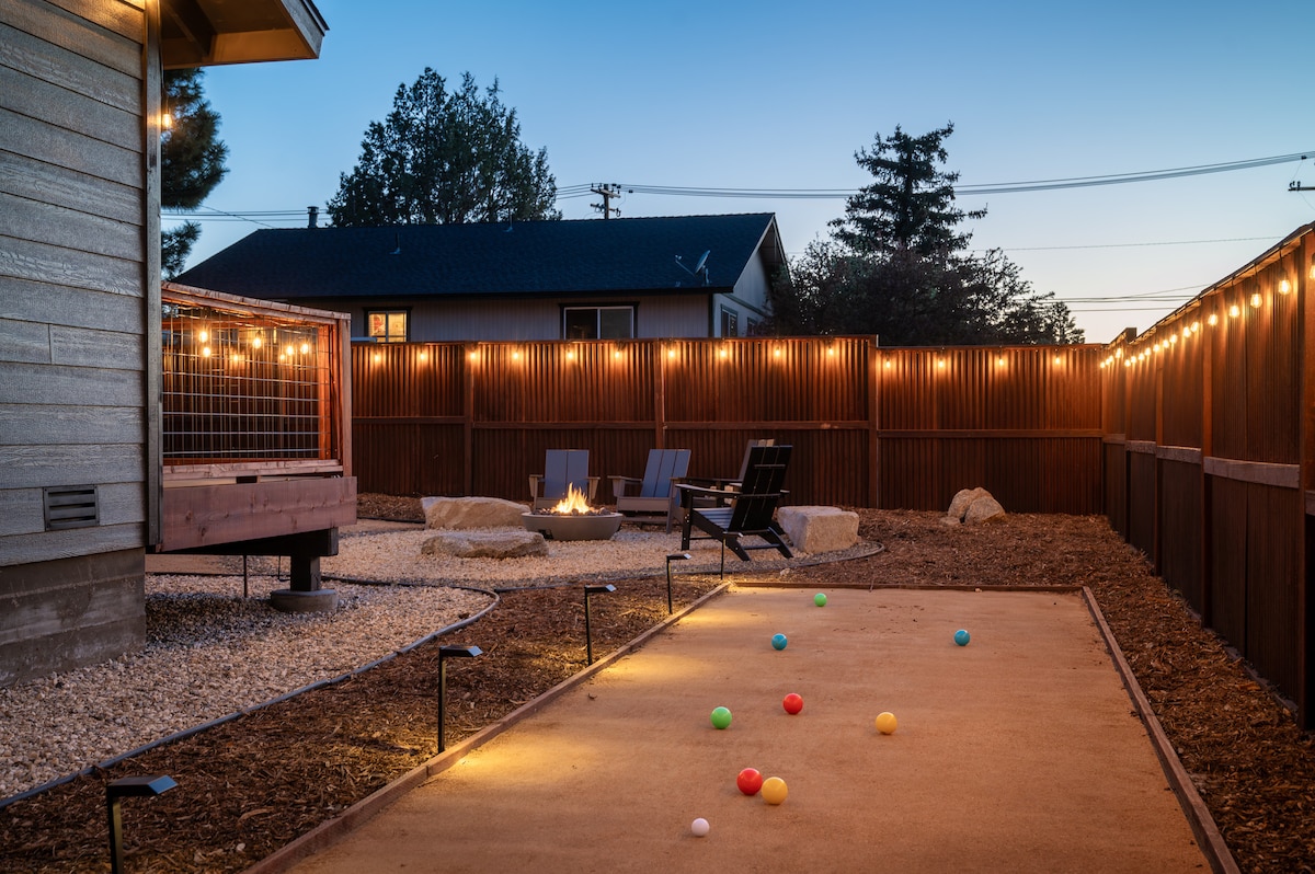 An outdoor space is showcased, featuring a fire pit surrounded by seating options. Pathway lights illuminate a bocce ball court, which is scattered with colorful balls. Soft lighting from string lights enhances the ambiance against the backdrop of a wooden fence.