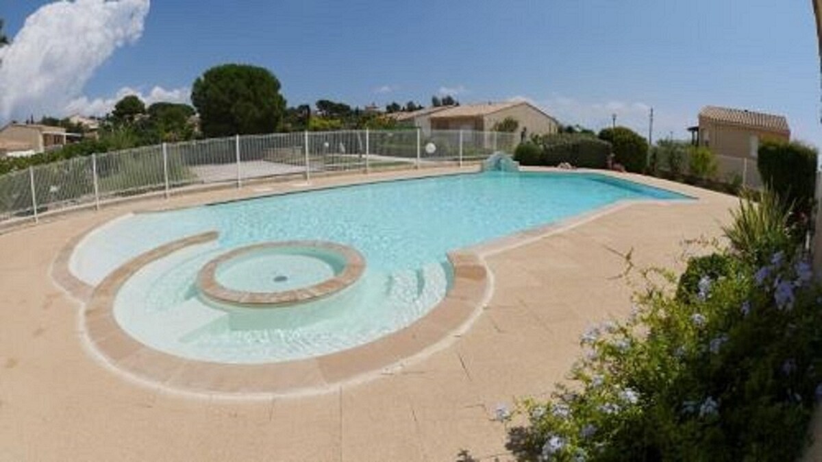 A swimming pool is prominently featured, surrounded by a neat fence. The pool includes a circular whirlpool section, with clear blue water shimmering under a bright sky. A landscaped area with greenery and flowers borders the pool, enhancing the outdoor setting.