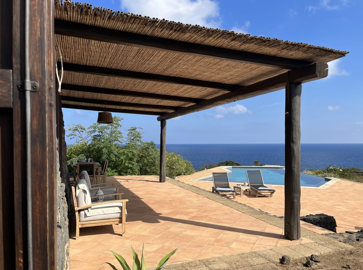 An outdoor terrace is shaded by a rustic wooden roof, featuring comfortable seating arrangements. Beyond the terrace, an infinity pool extends towards the sea, framed by a clear blue sky and greenery in the background, providing a sense of privacy and relaxation.