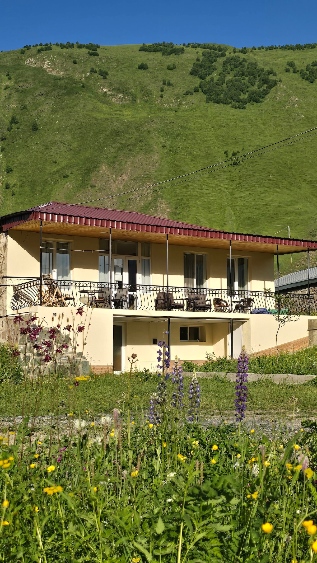 The two-story building features a spacious balcony lined with multiple seating options. Surrounding flowers and greenery add natural beauty, with a mountain backdrop creating a serene environment. The exterior is painted in soft tones, contributing to its relaxed and inviting presence.