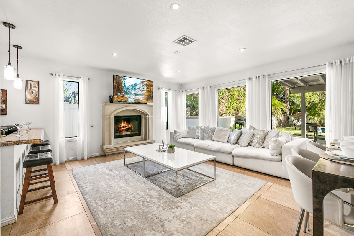 A spacious living room features a large sectional sofa adorned with soft cushions. A coffee table sits centrally, while a fireplace adds warmth. Natural light fills the room through multiple windows, leading to an outdoor space visible in the background, enhancing the inviting environment.