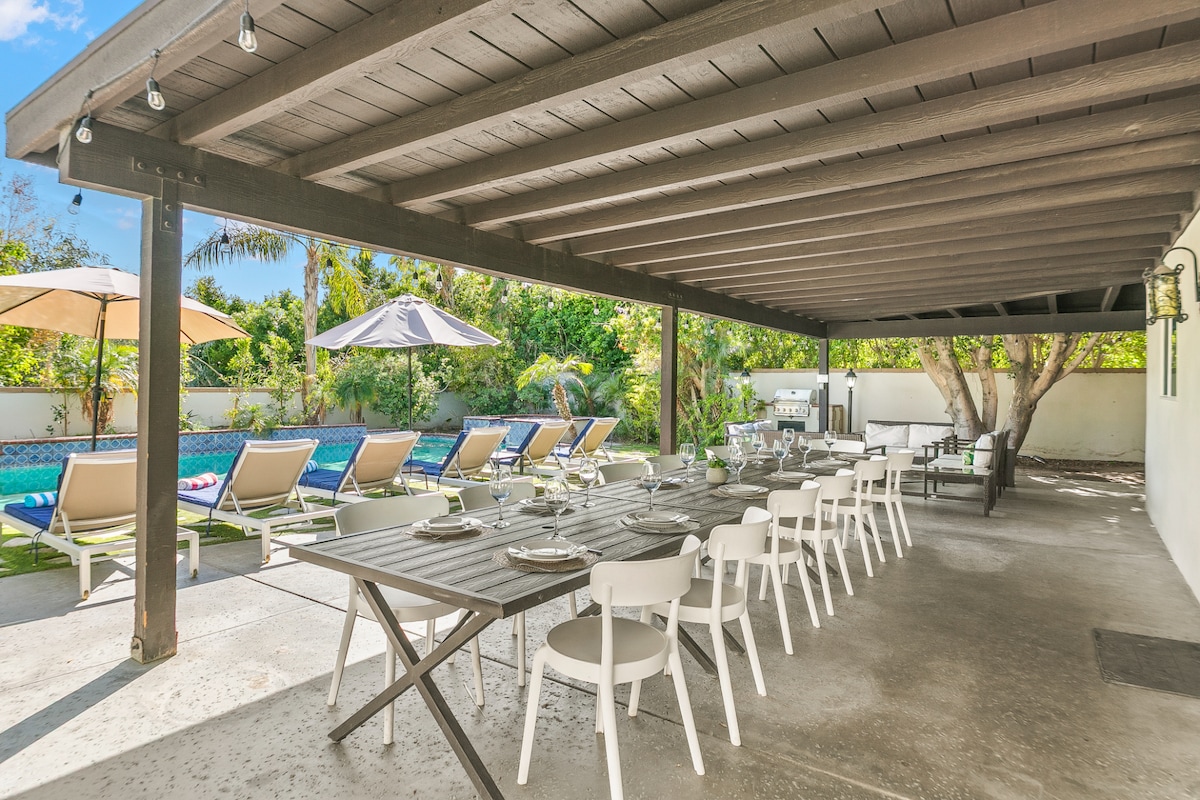A shaded outdoor dining area is equipped with a long wooden table and white chairs, positioned next to a private pool. Lounge chairs line the poolside beneath large umbrellas, surrounded by lush greenery, creating a serene setting for relaxation and gatherings.