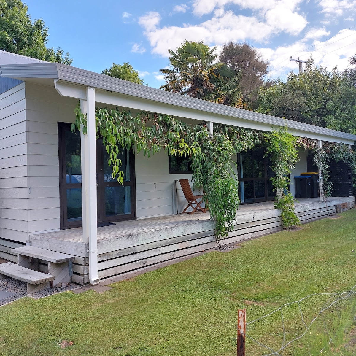 A single-story cottage presents a covered wooden deck surrounded by greenery. Two wooden chairs are positioned on the deck, while large windows invite natural light into the interior. A spacious lawn leads to the cottage's entrance, creating a serene outdoor environment.