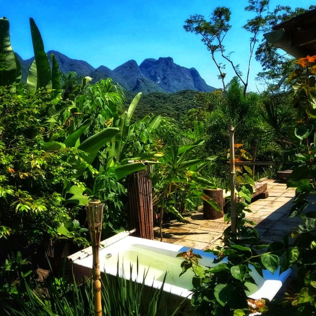 A vintage-style bathtub is nestled amidst lush greenery, surrounded by tropical plants. The backdrop features majestic mountains under a clear blue sky, creating a serene and natural environment.