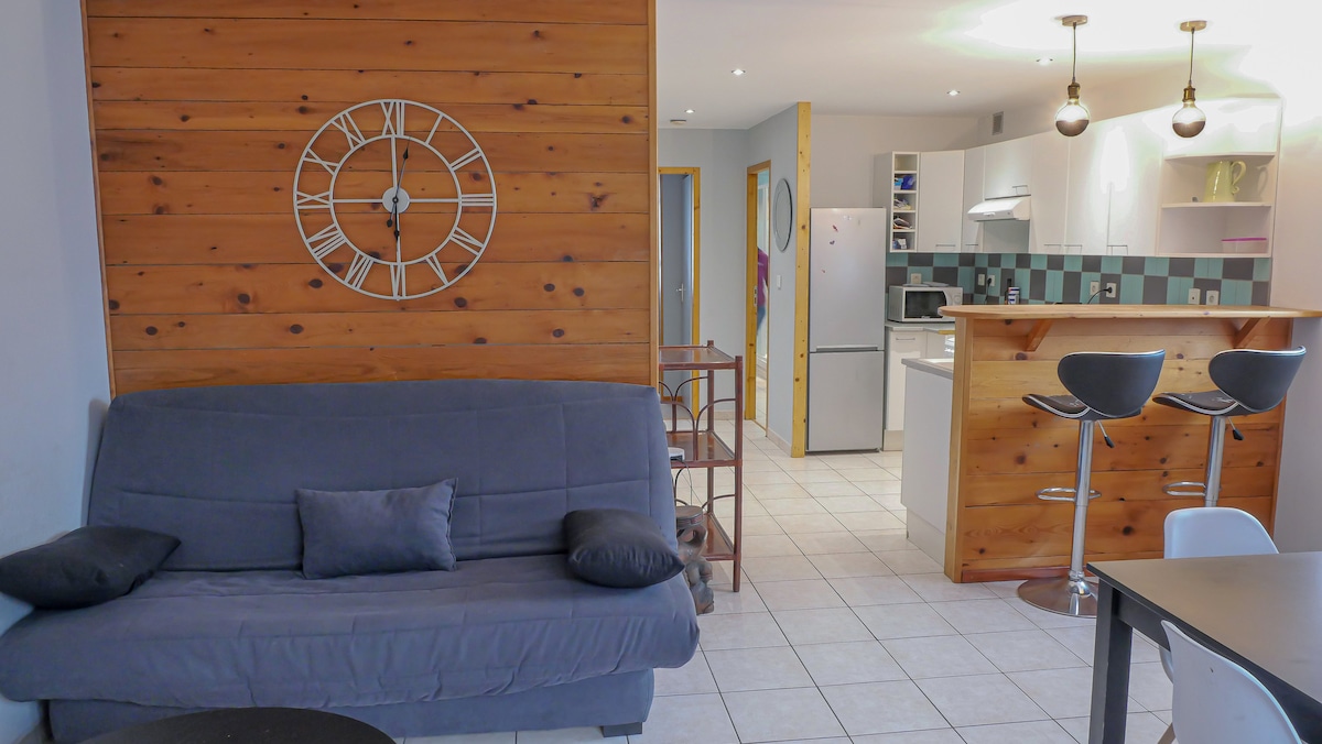 An open living space displays a gray sofa bed and a wooden feature wall with a large clock. The kitchen area is partially visible, featuring modern appliances and a breakfast bar with two high stools, enhancing the communal atmosphere.