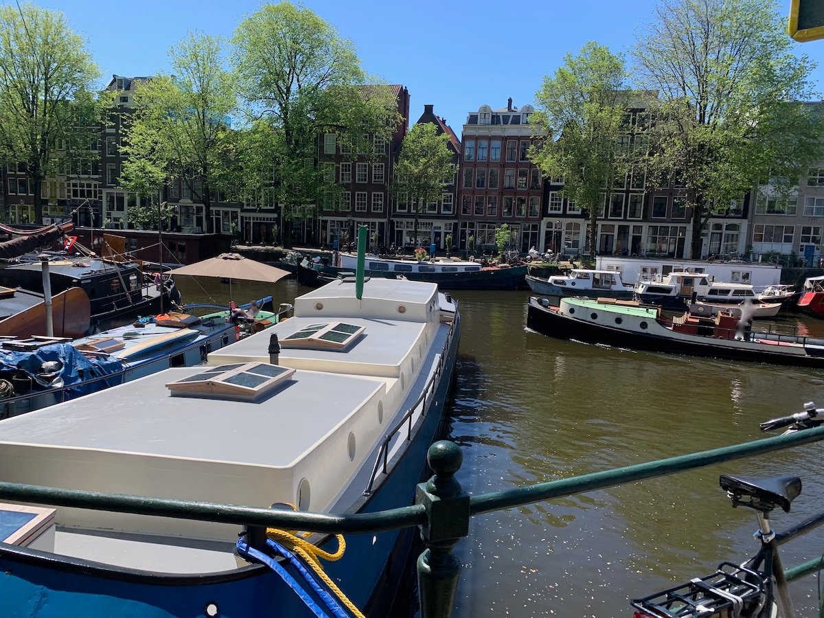 The image features a scenic view of a waterway lined with houseboats. Lush green trees provide shade along the banks, while traditional Dutch buildings are visible in the background. Several boats are anchored in the water, hinting at a vibrant community atmosphere.