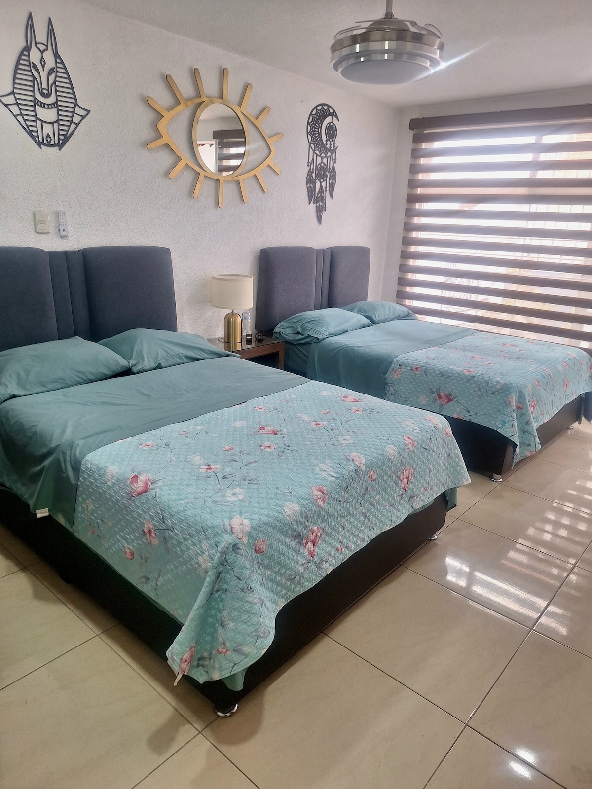 A well-lit room features two spacious beds with aqua-colored bedding adorned with floral patterns. The walls exhibit decorative accents, including a round mirror and wall art, while natural light filters through classic horizontal blinds.