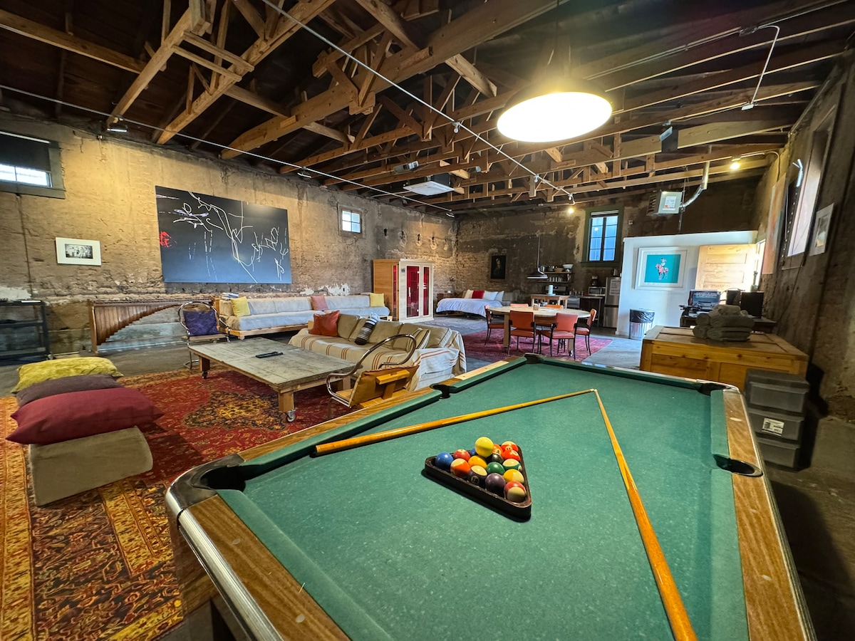 A spacious room features a commercial pool table in the foreground, surrounded by multiple seating areas including couches and day beds. Hardwood beams overhead create an open atmosphere, while artwork adorns the walls. A large window lets in natural light, enhancing the room's inviting feel.