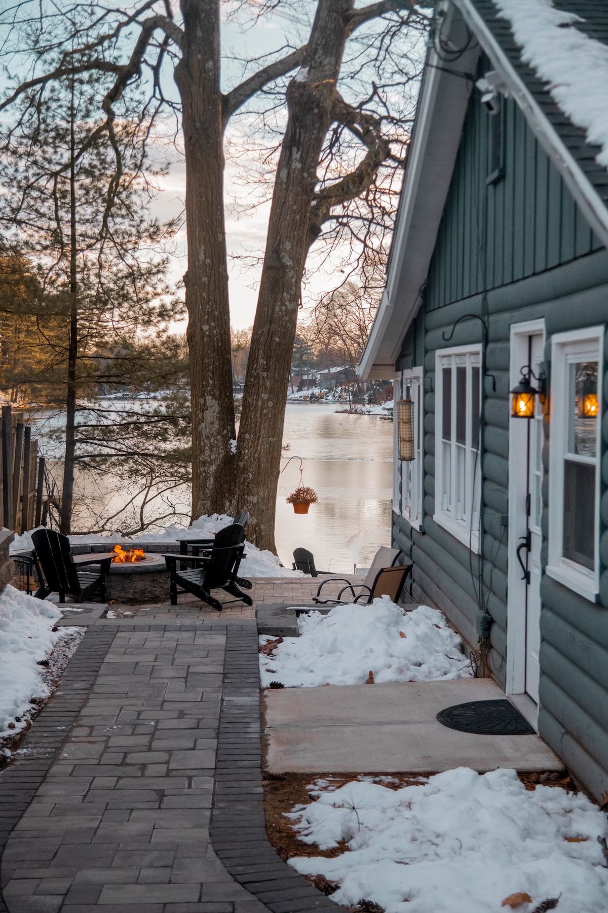 A stone pathway leads to a fire pit area with black outdoor chairs, surrounded by snow. The charming exterior of the cottage is seen, with a view of the lake and trees in the background.