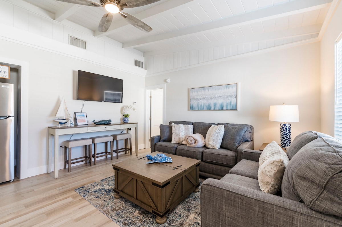 A cozy living area features two gray sofas flanking a coffee table. Light wood flooring complements the white ceiling with exposed beams. A flat-screen TV is mounted on the wall across from a wooden dining table with four bar stools. Natural light fills the space through a large window.
