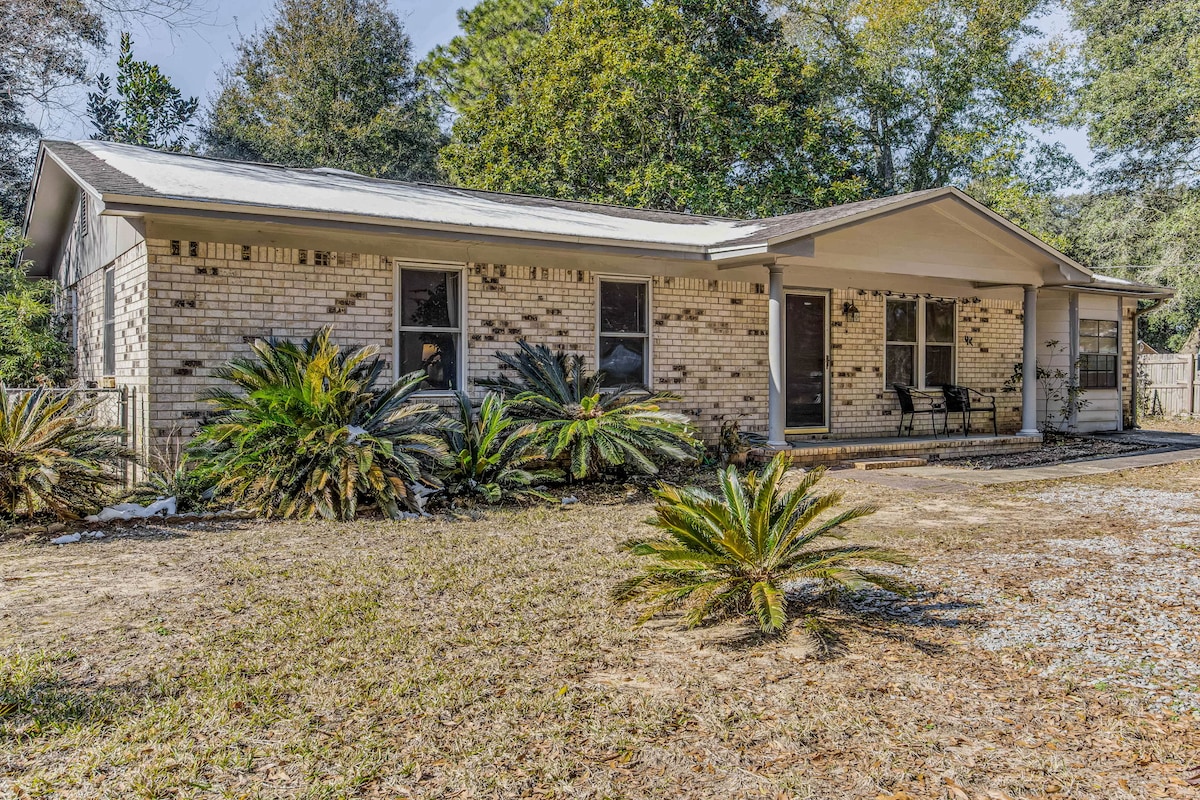 The exterior of the home features a single-story structure with a brick facade surrounded by lush greenery. Two chairs are positioned on the porch, and large palm plants line the front, creating a spacious outdoor area. Natural light enhances the inviting appearance of the property.