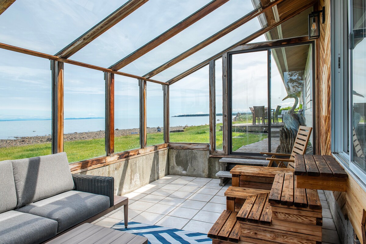 A sunlit enclosed porch is depicted with a comfortable grey sofa and wooden tables. Large glass panels provide unobstructed views of the ocean beyond. The flooring features white tiles, while a blue and white patterned rug adds a subtle touch of color.