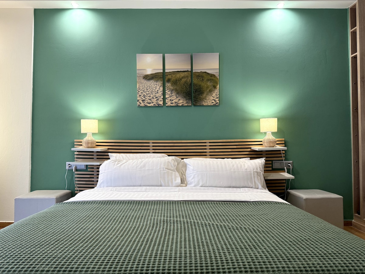 A spacious sleeping area features a wooden headboard with a soft bedding arrangement. Two bedside lamps illuminate the space, while a triptych artwork depicting a beach scene adds a calming touch. The walls are painted in a soothing green hue.