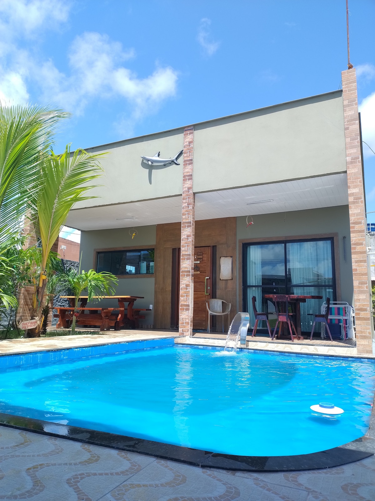 A private outdoor area features a pool with bright blue water, surrounded by tiled flooring and tropical plants. A wooden dining table is seen under a covered area, and lounge chairs are positioned nearby. Bright sunlight and a clear sky enhance the inviting space.